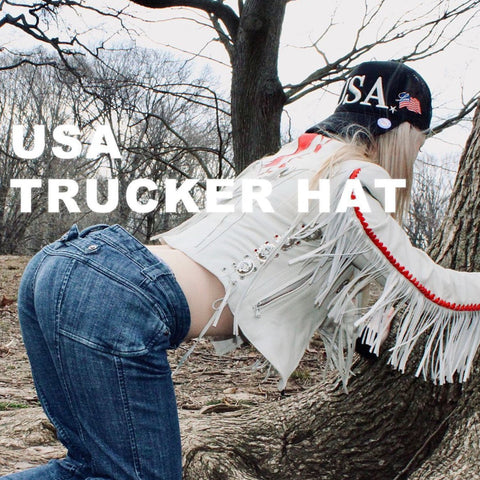 A person wearing a black 'USA TRUCKER HAT' with a white top and blue jeans, posing in front of a tree.