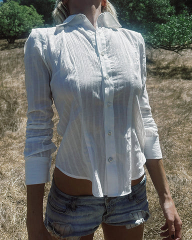 A woman wearing a white long-sleeve button-up shirt with subtle vertical texture and light blue denim shorts, standing outdoors in a dry grassy field.